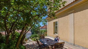 An outdoor patio with a table and chairs, surrounded by lush greenery and a building with a tiled roof.