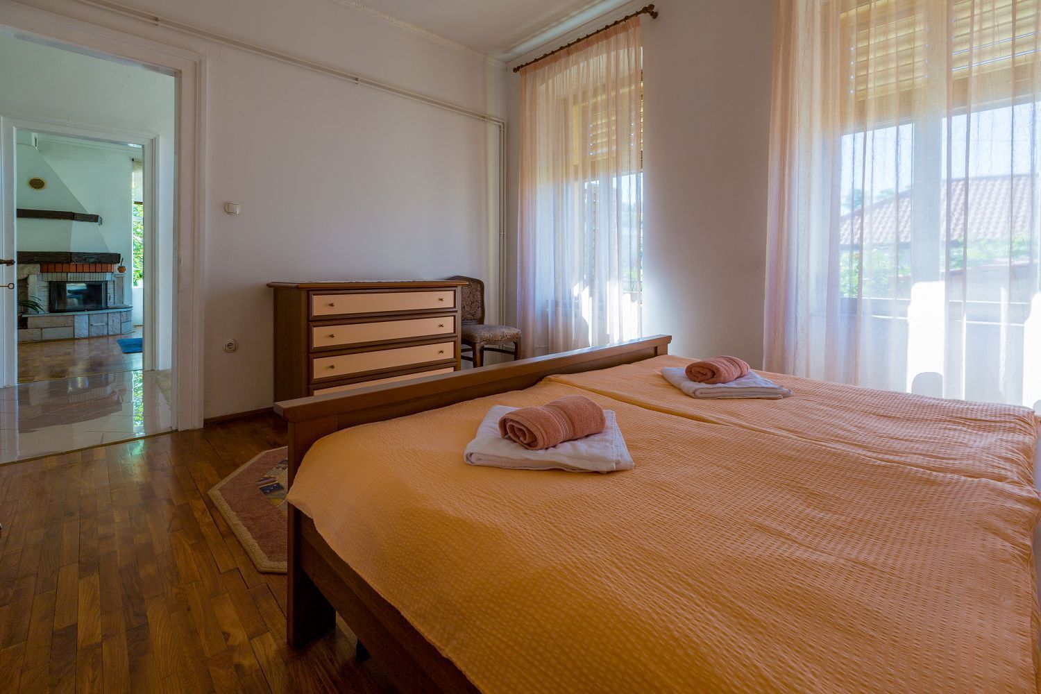 Habitación de hotel con una cama grande, un aparador de madera y vistas al exterior a través de cortinas traslúcidas.