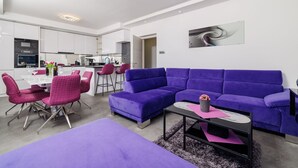 Ein modernes Wohnzimmer mit einem violetten Ecksofa, einem Essbereich mit rosafarbenen Stühlen und einer Küche im Hintergrund.