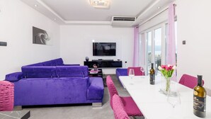 Ein modernes Wohnzimmer mit einem violetten Sofa, einem Esstisch mit rosa Stühlen, einem Fernseher und Meerblick.