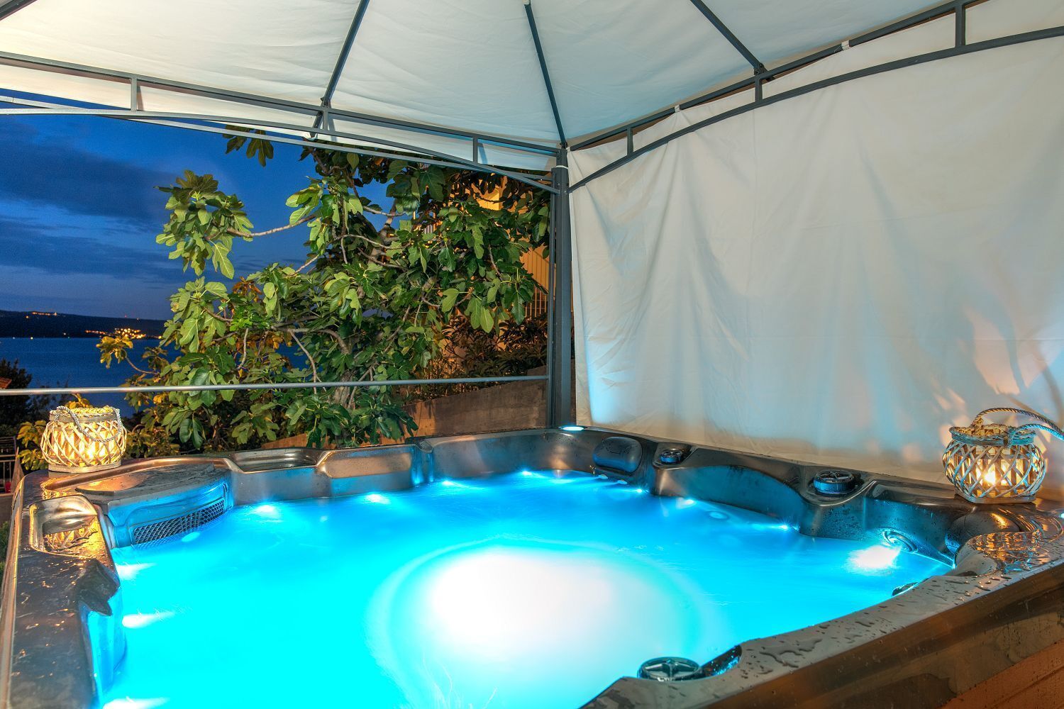 Un jacuzzi al aire libre con vista al mar de noche, rodeado de follaje bajo una cubierta blanca.