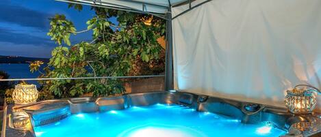 Un jacuzzi al aire libre con vista al mar de noche, rodeado de follaje bajo una cubierta blanca.
