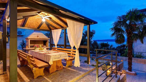 An outdoor dining area with a covered patio, wooden benches, a table set with plates and candles, and a view of the sea. - Stunning flat 150 metres to the beach with jacuzzi, sea view, pool, pool table (Dramalj)