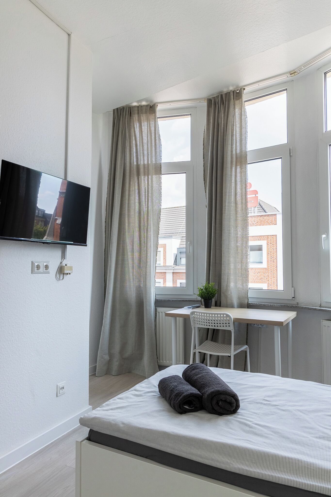 A bedroom with a bed, a desk, a chair, a TV mounted on the wall, and a window with curtains.