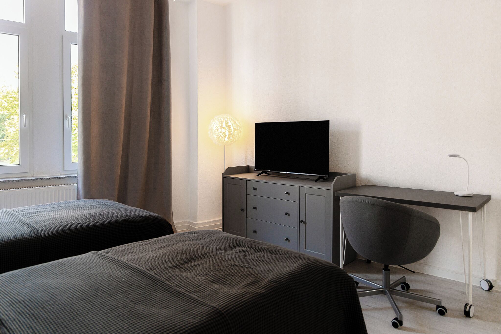 A bedroom with two beds, a desk with a chair, a television, and a lamp.