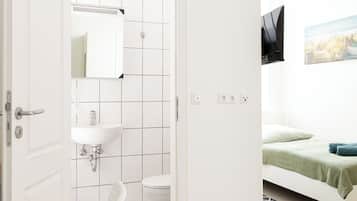 Una habitación moderna y minimalista con una puerta blanca que da al baño, una cama con colcha verde y un televisor montado en la pared.