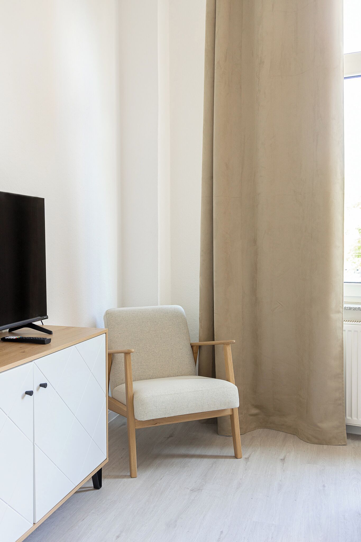 A room with a television on a wooden stand, a white cabinet, a beige chair, and a large beige curtain.
