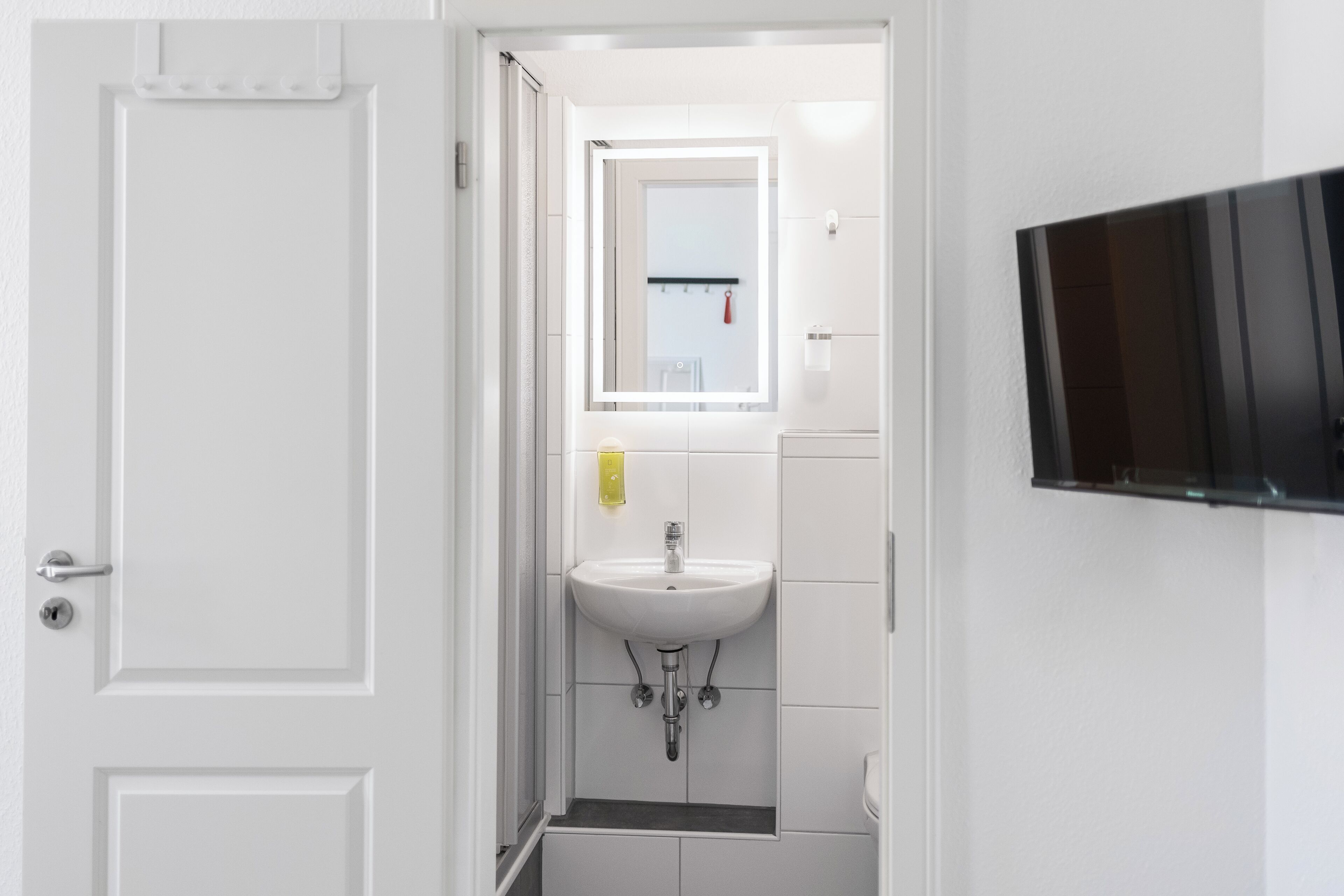 Un baño con un lavamanos blanco, un espejo y un televisor negro montado en la pared.