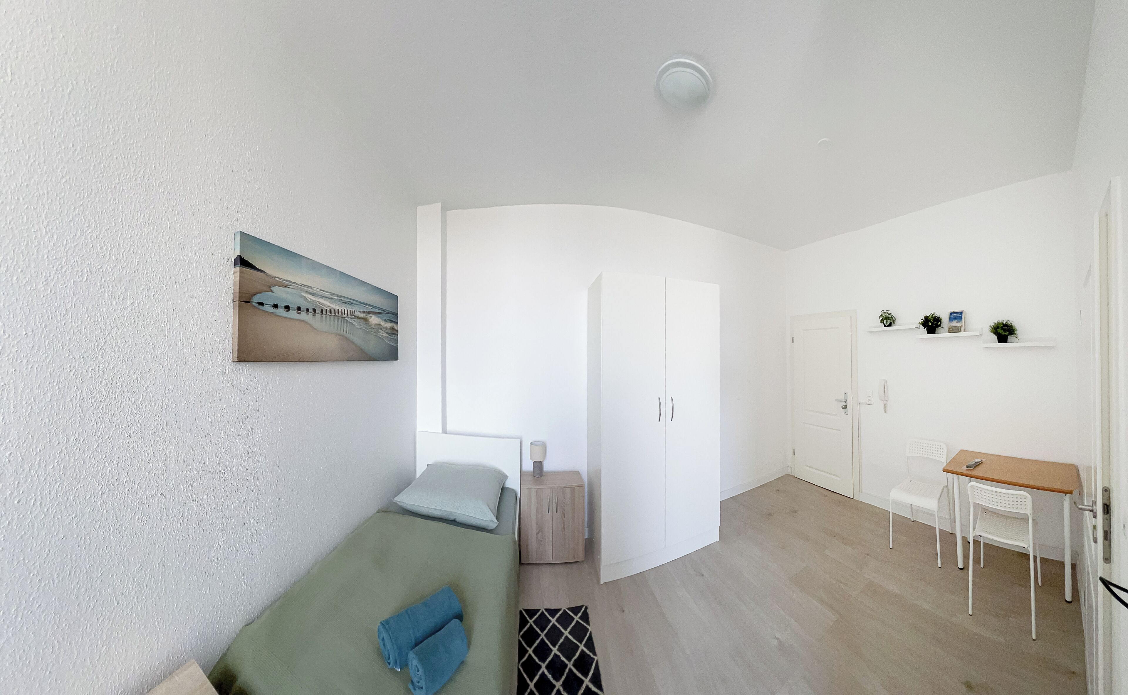 Quarto compacto com uma cama, uma escrivaninha e uma porta que dá para outro quarto.