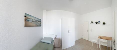 Quarto compacto com uma cama, uma escrivaninha e uma porta que dá para outro quarto.