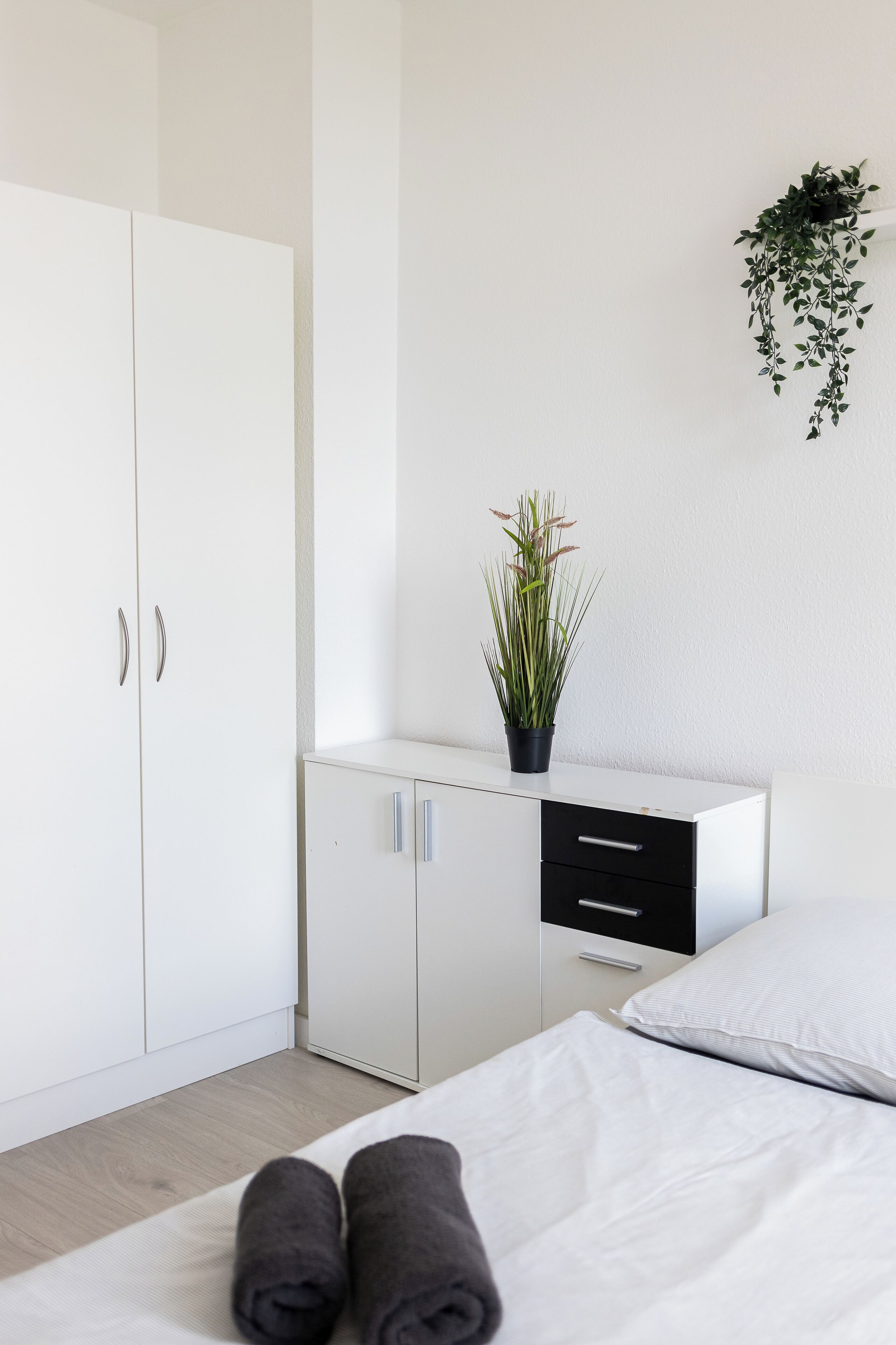 A minimalist bedroom with a bed, a nightstand, a wardrobe, and a hanging plant.