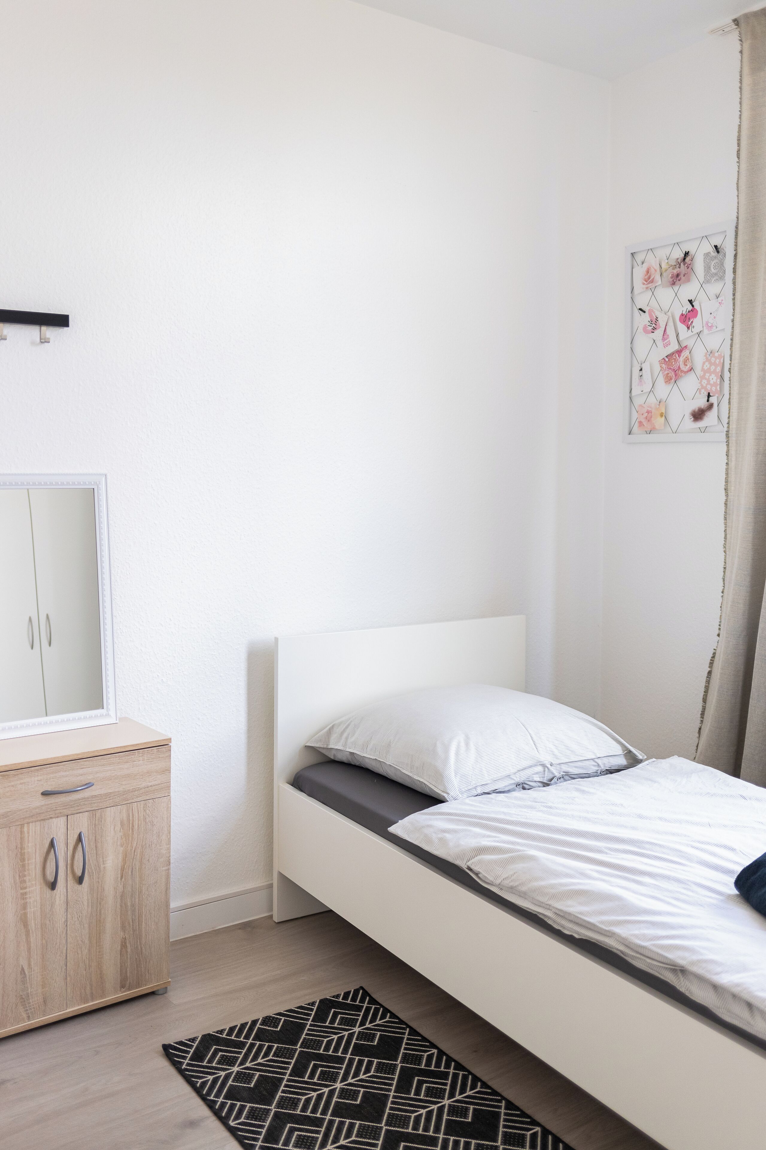 Ein Schlafzimmer mit einem Einzelbett, einem Holzschrank, einem Spiegel und einem Teppich mit geometrischem Muster.