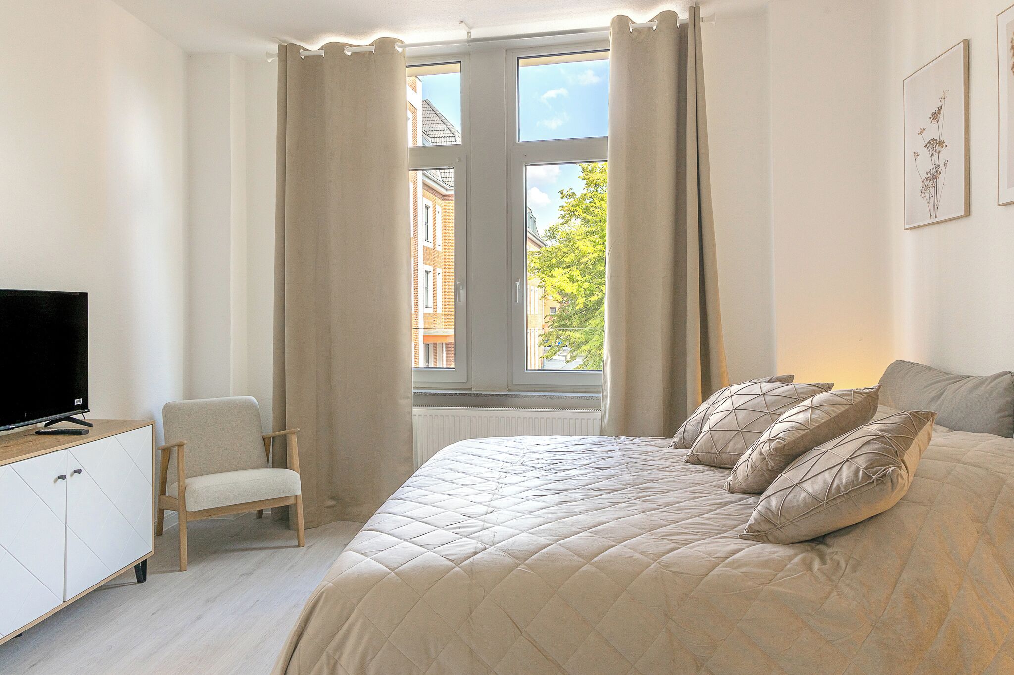 Ein Schlafzimmer mit Bett, Stuhl, Fernseher und einem Fenster mit Vorhängen.