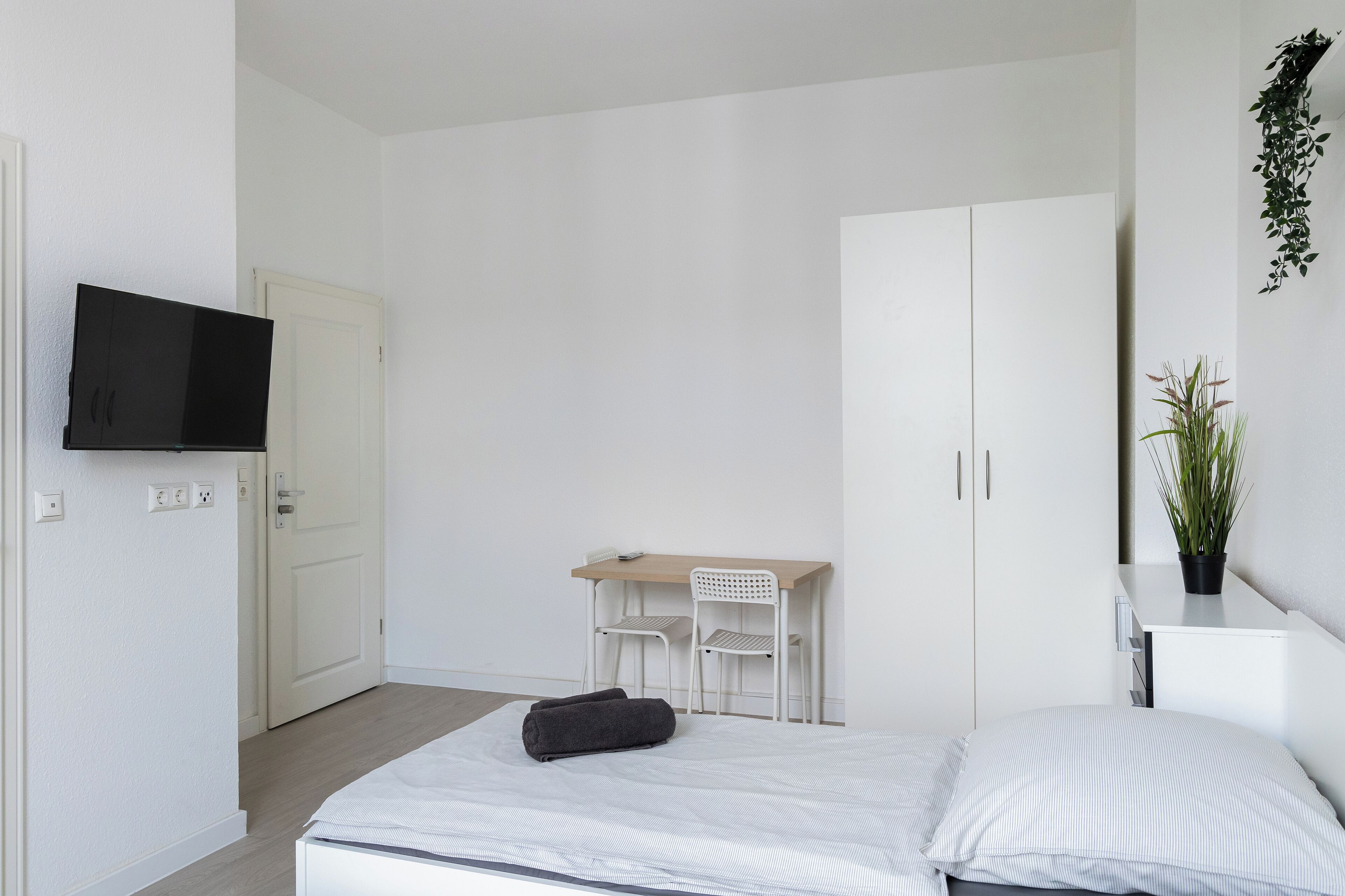 Een minimalistische slaapkamer met een bed, een bureau, twee stoelen, een televisie en een potplant.