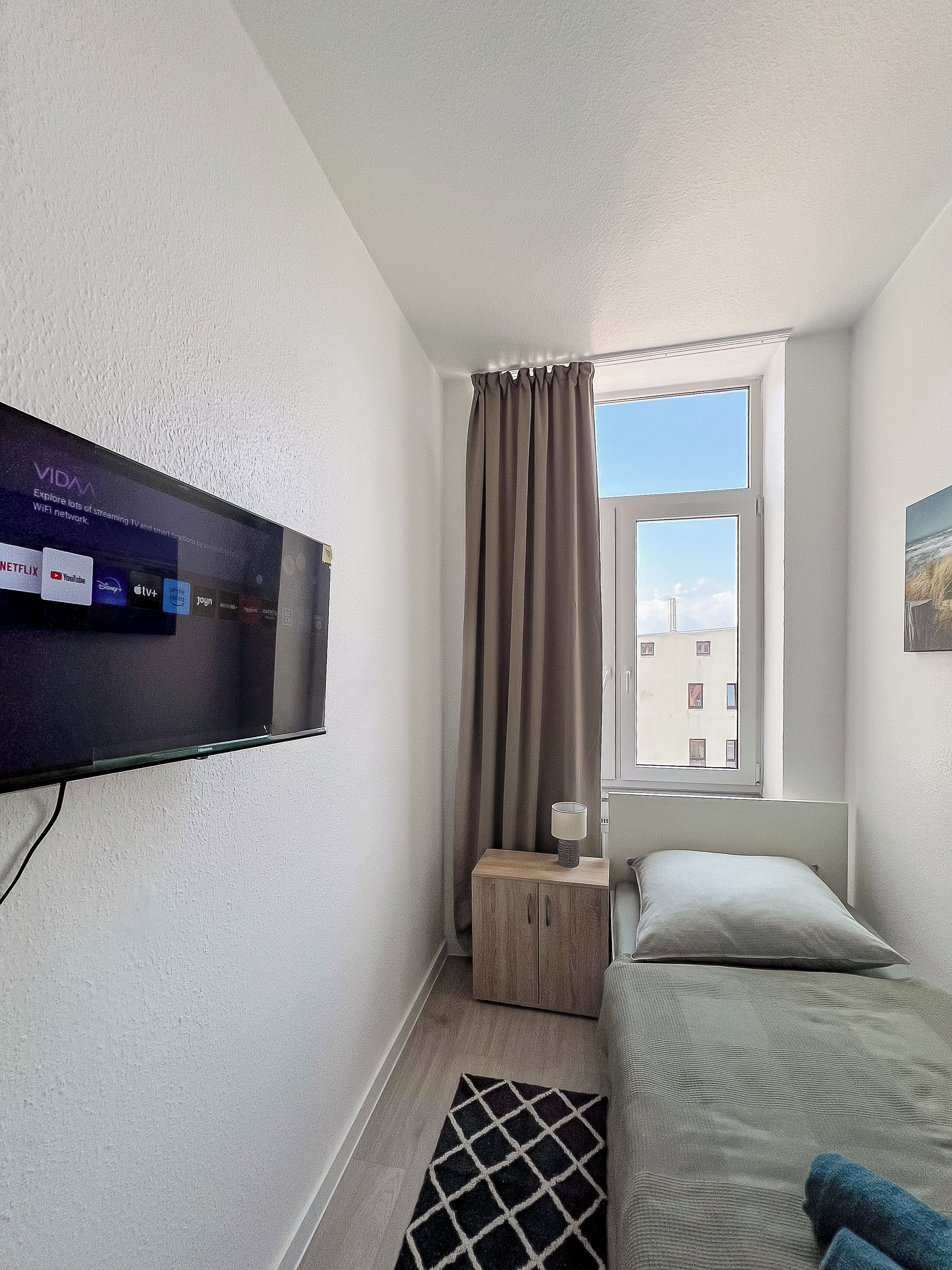 Un dormitorio con televisión montada en la pared, ventana con cortinas, mesita de noche con lámpara y cama con almohada.
