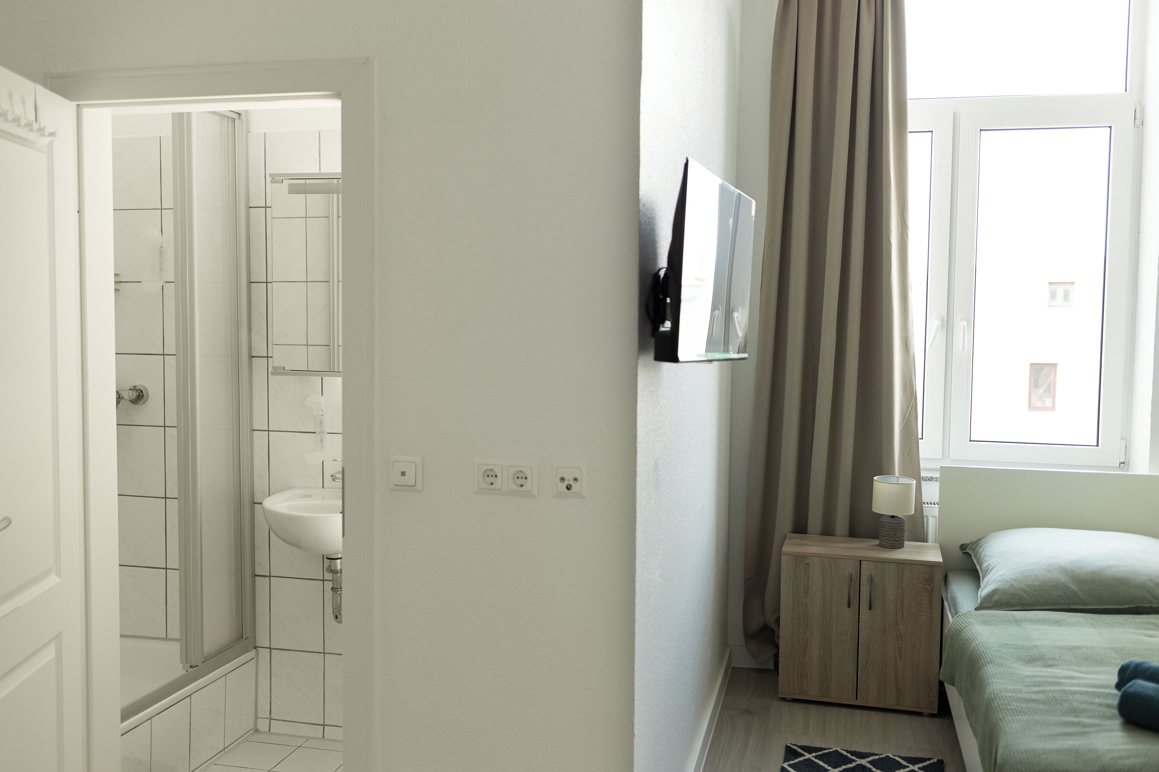 A hotel room with a shower, a bedside table with a lamp, a bed with a green bedspread, and a window with curtains.