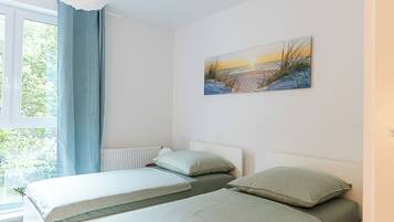 A bedroom with two beds, a window with blue curtains, and a painting of a sunset on the wall.
