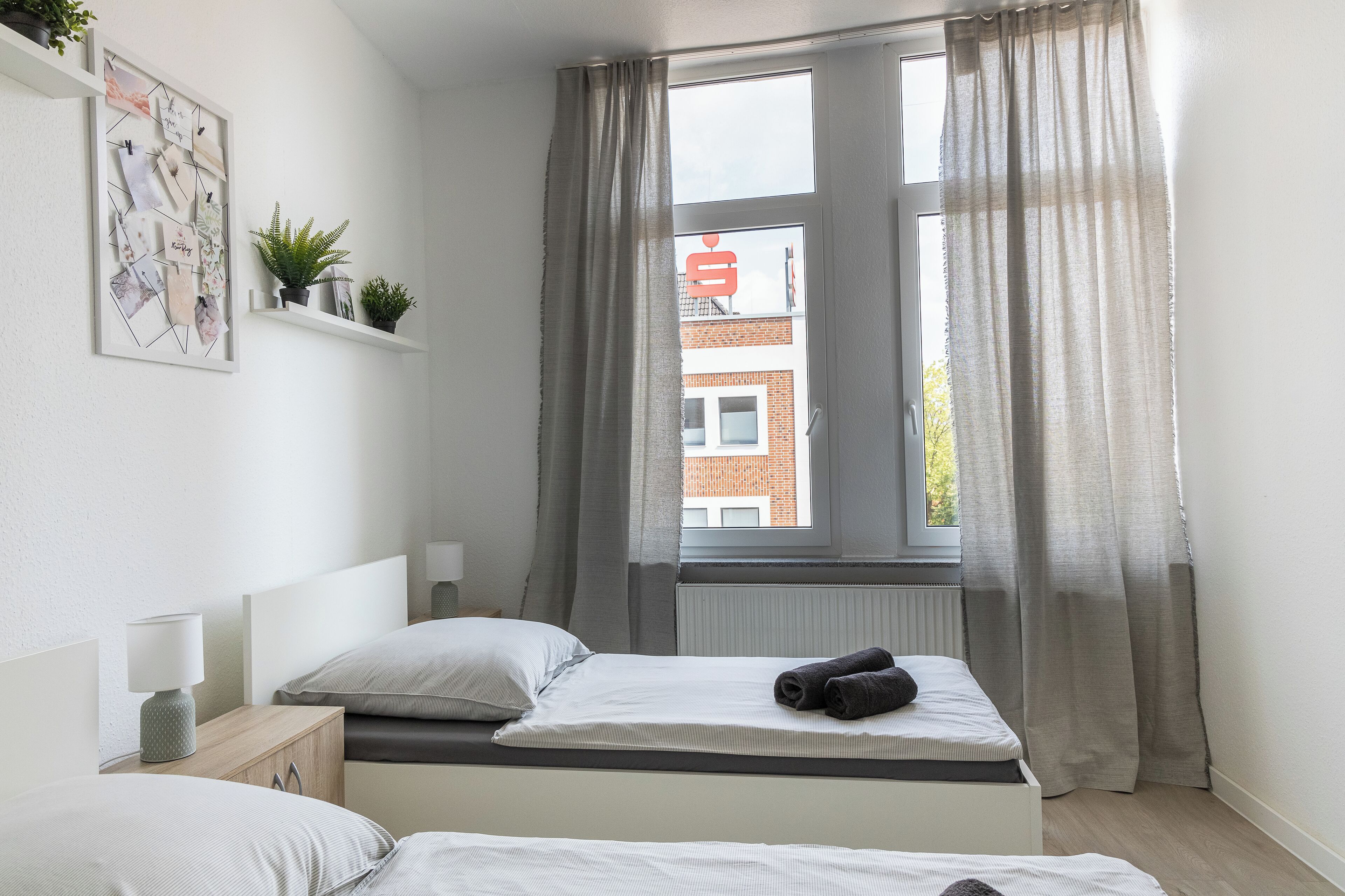 A bedroom with two beds, a window with curtains, a wall-mounted shelf with plants, and a framed picture.