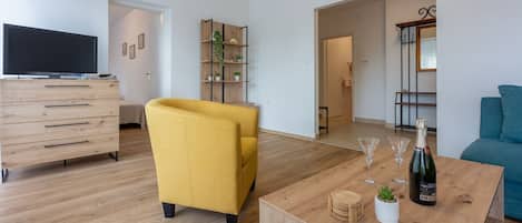 Een moderne woonkamer met houten vloer, een gele fauteuil, een houten salontafel met glazen en een fles, en een blauwe bank.
