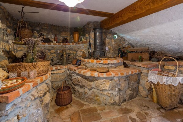 A rustic stone interior with a wood beam ceiling, stone walls, and a central chimney.