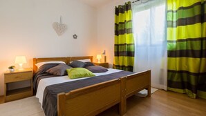 Una cama de madera con colchón blanco, dos mesitas de noche con lámparas, una decoración en la pared con forma de corazón y una ventana con cortinas a rayas.