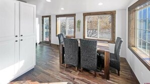 A dining area with a wooden table and chairs, white cabinets, and large windows with wooden frames. - Colorado Willow House (Cortez)