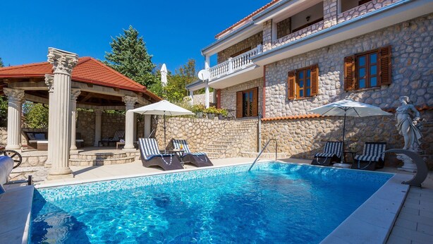 A swimming pool with lounge chairs and umbrellas, stone walls, and a covered seating area with columns.