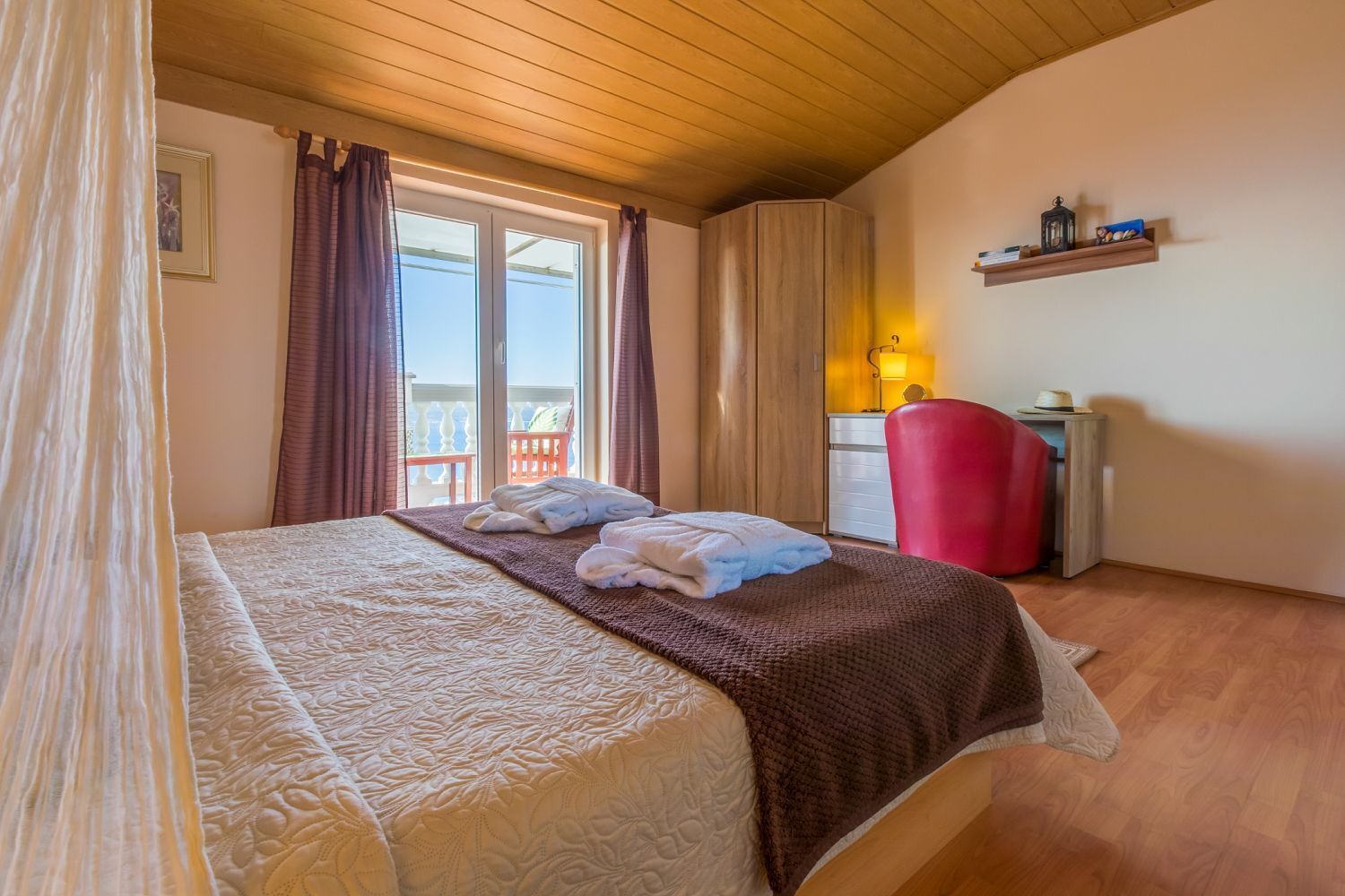 Una camera da letto con un letto, una poltrona rossa, un comodino, una finestra con tende e una mensola con oggetti.