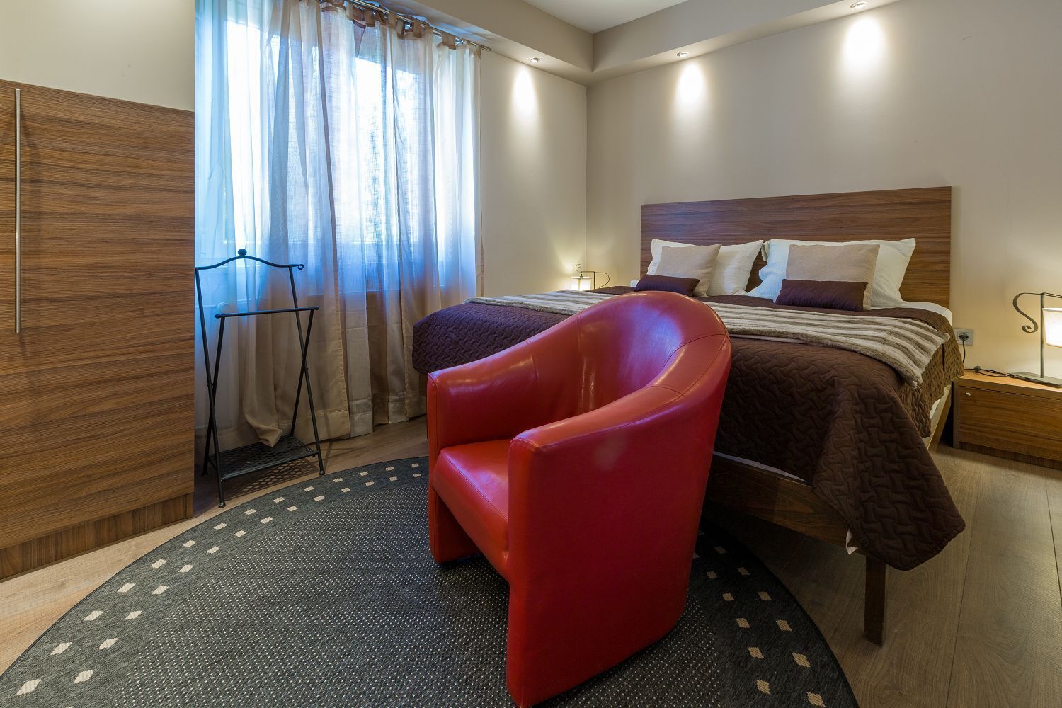 A hotel room with a bed, a red chair, a standing lamp, and a window with curtains.