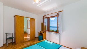 Une chambre avec une armoire en bois, un lit simple avec une literie bleue, une chaise, une petite table et une fenêtre donnant sur la mer.