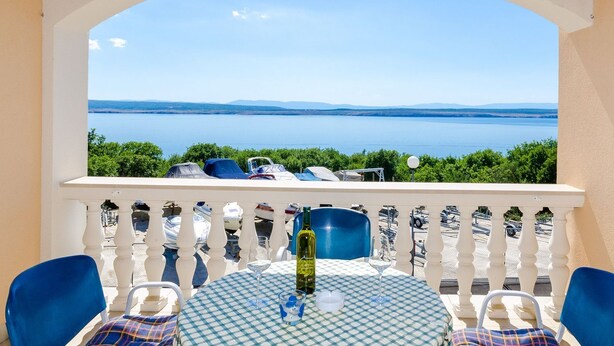 Un balcon avec une nappe à carreaux, une balustrade blanche et une vue sur une étendue d’eau et la verdure.