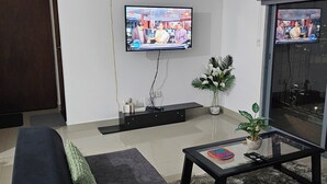 Una sala moderna con un televisor de pantalla plana montado en la pared, un sofá negro, una mesa de centro de vidrio con controles remotos y una planta en maceta.
