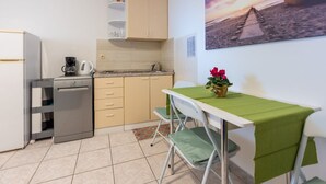 Eine kleine Küche mit Kühlschrank, Geschirrspüler und einer Kaffeemaschine. Ein Esstisch mit grüner Tischdecke und ein Stuhl stehen bereit. Ein Bild mit Strandmotiv ist an der Wand angebracht.