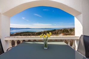 A balcony with a table and chairs, a vase with yellow flowers, and a scenic view of a coastline and mountains.