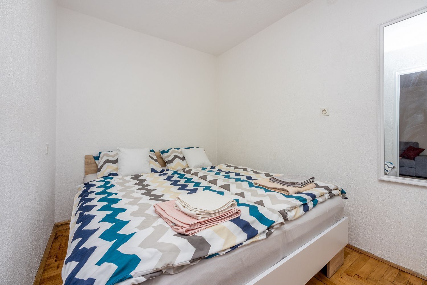 Una cama pequeña y ordenada con una colcha estampada y almohadas, suelo de madera y una ventana que da a un sofá.