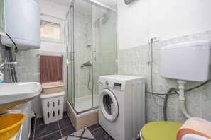 A compact bathroom with a glass-enclosed shower, a washing machine, a toilet, and a sink.