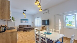 Ein modernes Wohnzimmer mit Sofa, Fernseher, Esstisch und Stühlen. Es gibt eine Küchenzeile mit Wasserkocher und einer Pflanze.