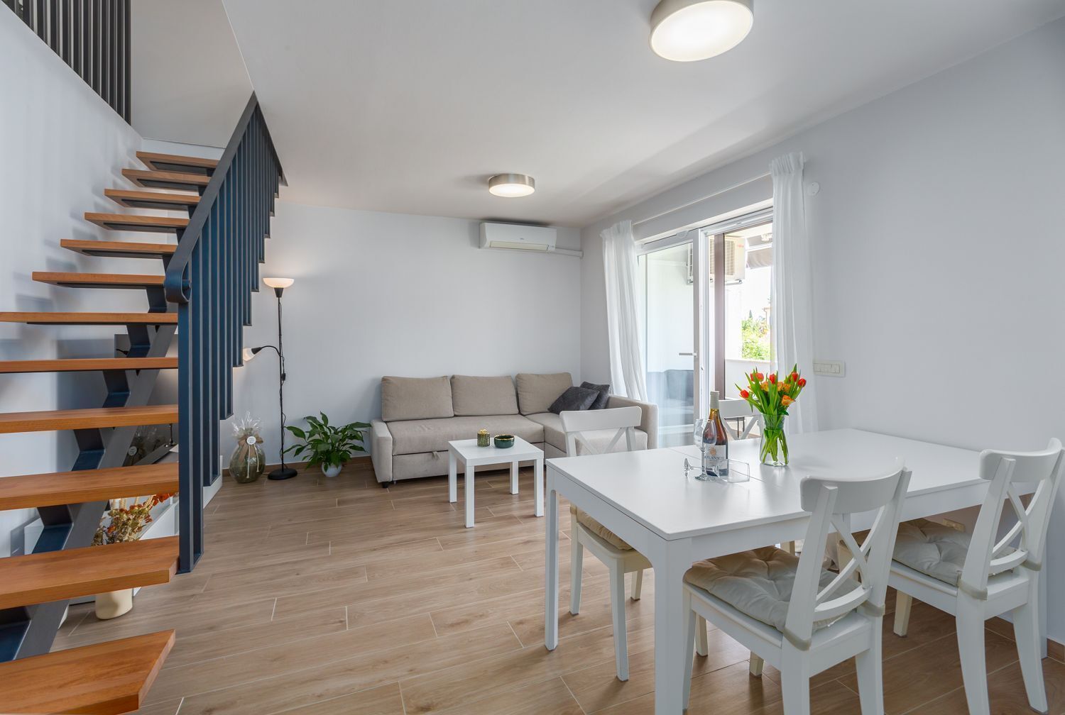Ett modernt vardagsrum med en trappa, en soffa, ett matbord och en krukväxt.
