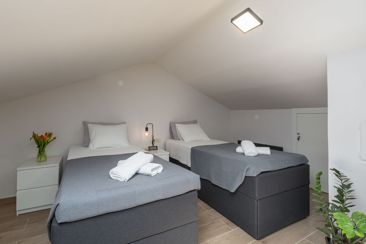 A modern, well-lit room with two beds, a bedside table with a lamp, a small plant, and a vase of flowers.