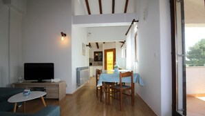 Un salon avec une télévision, une petite table et un canapé. Il y a également un coin repas avec une table et des chaises. La pièce est dotée de parquets en bois et de murs blancs.