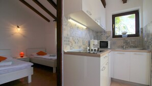 Une chambre compacte avec un lit simple et une table de chevet, une petite kitchenette équipée d’un four à micro-ondes et d’une machine à café, et une fenêtre donnant sur des palmiers.