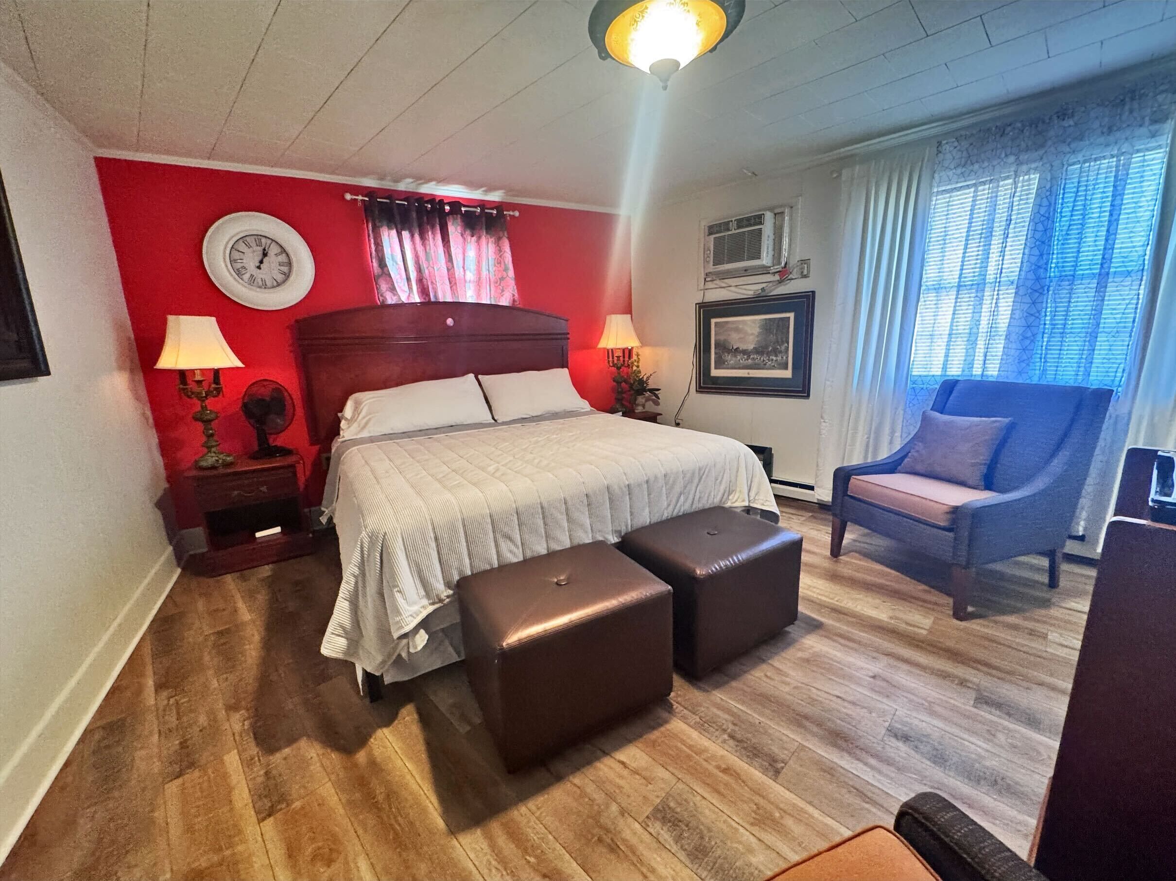 Un dormitorio con una cama, dos mesitas de noche, una silla, una ventana con cortinas y un reloj de pared.