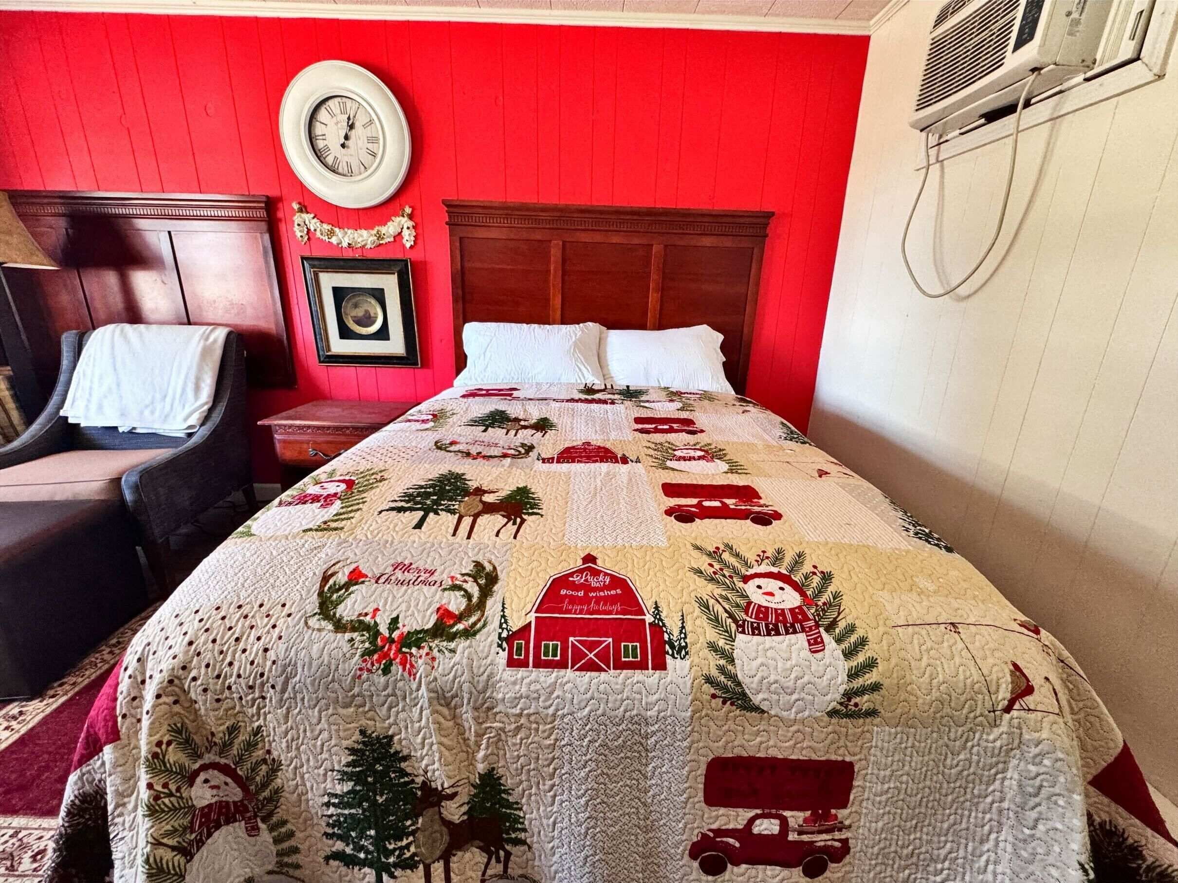Habitación de hotel con pared roja, cabecera de madera, cama con colcha navideña, un reloj y un cuadro enmarcado.