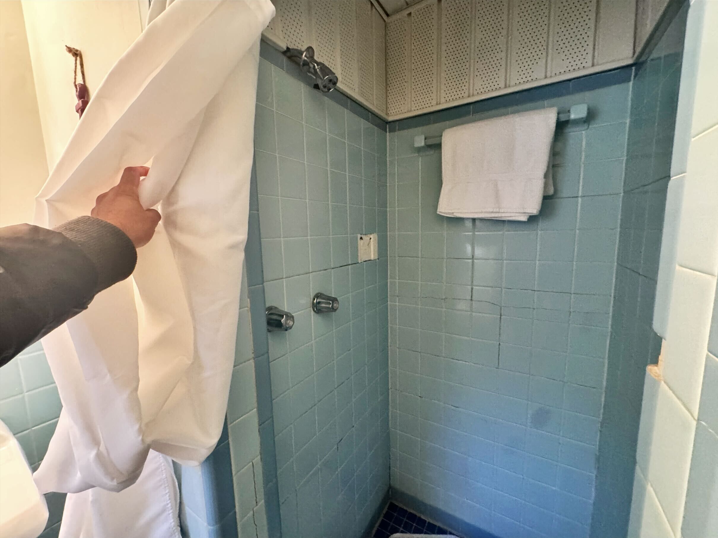 Un baño con paredes de azulejos azules, una toalla blanca colgada en un perchero y una cortina de ducha que se está acomodando.