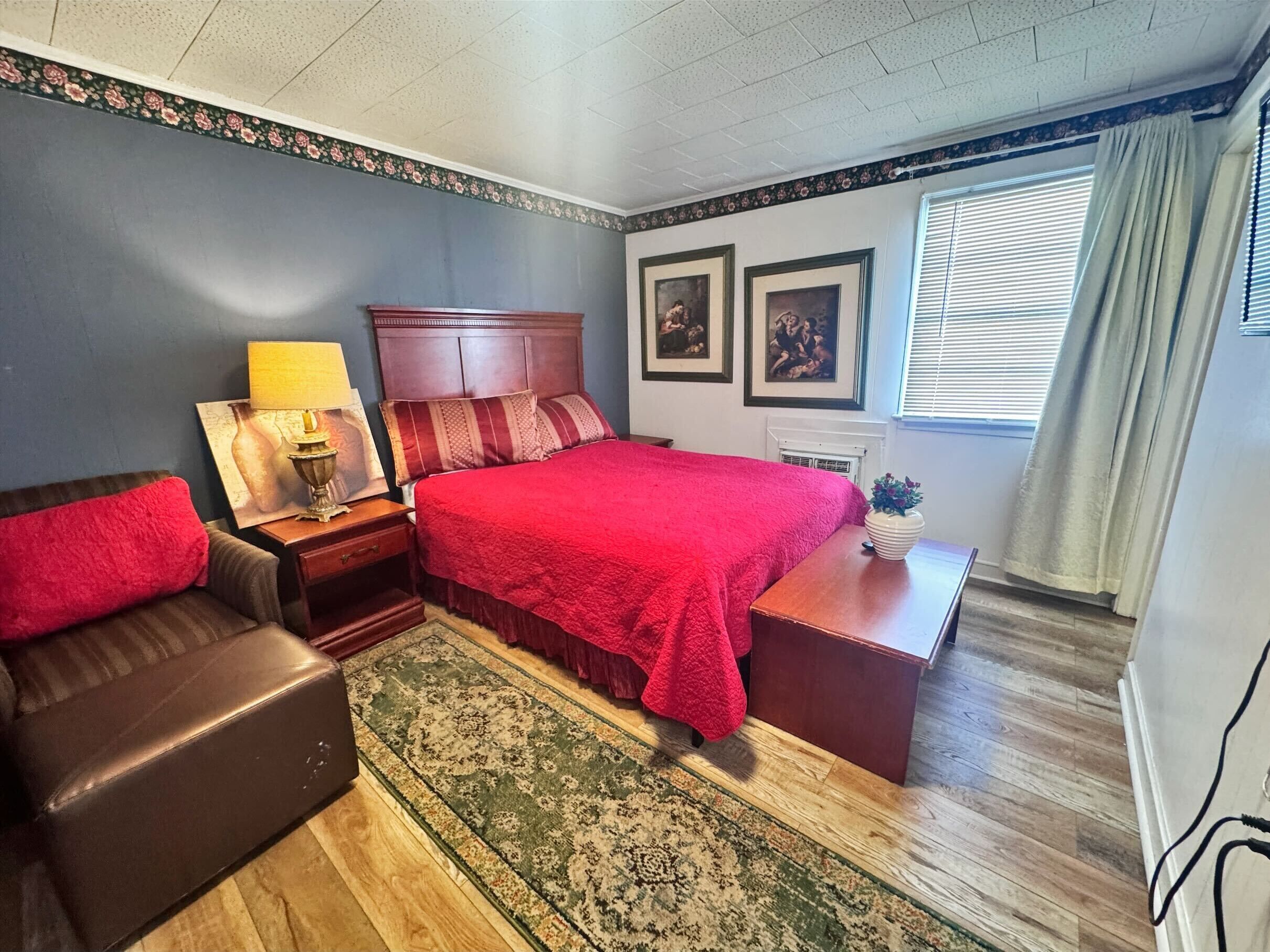 Un dormitorio con cama, mesita de noche, lámpara, silla, escritorio, ventana con cortinas y pared con cuadros.