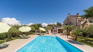 A rectangular swimming pool with clear blue water, surrounded by a paved deck. There are lounge chairs and umbrellas along the poolside, and a building with balconies in the background. - Idyllic flat just 250 metres from the beach with pool, sea view, barbecue area, loungers (Porec)