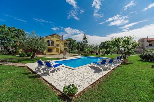 Une piscine entourée de chaises longues, une terrasse en pierre et, au fond, une maison jaune.