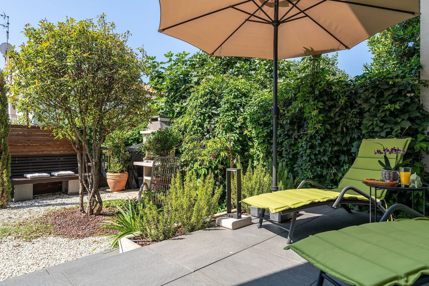 Un'area patio con una poltrona verde, un tavolo con un bicchiere di succo d'arancia e un ombrellone che offre ombra.
