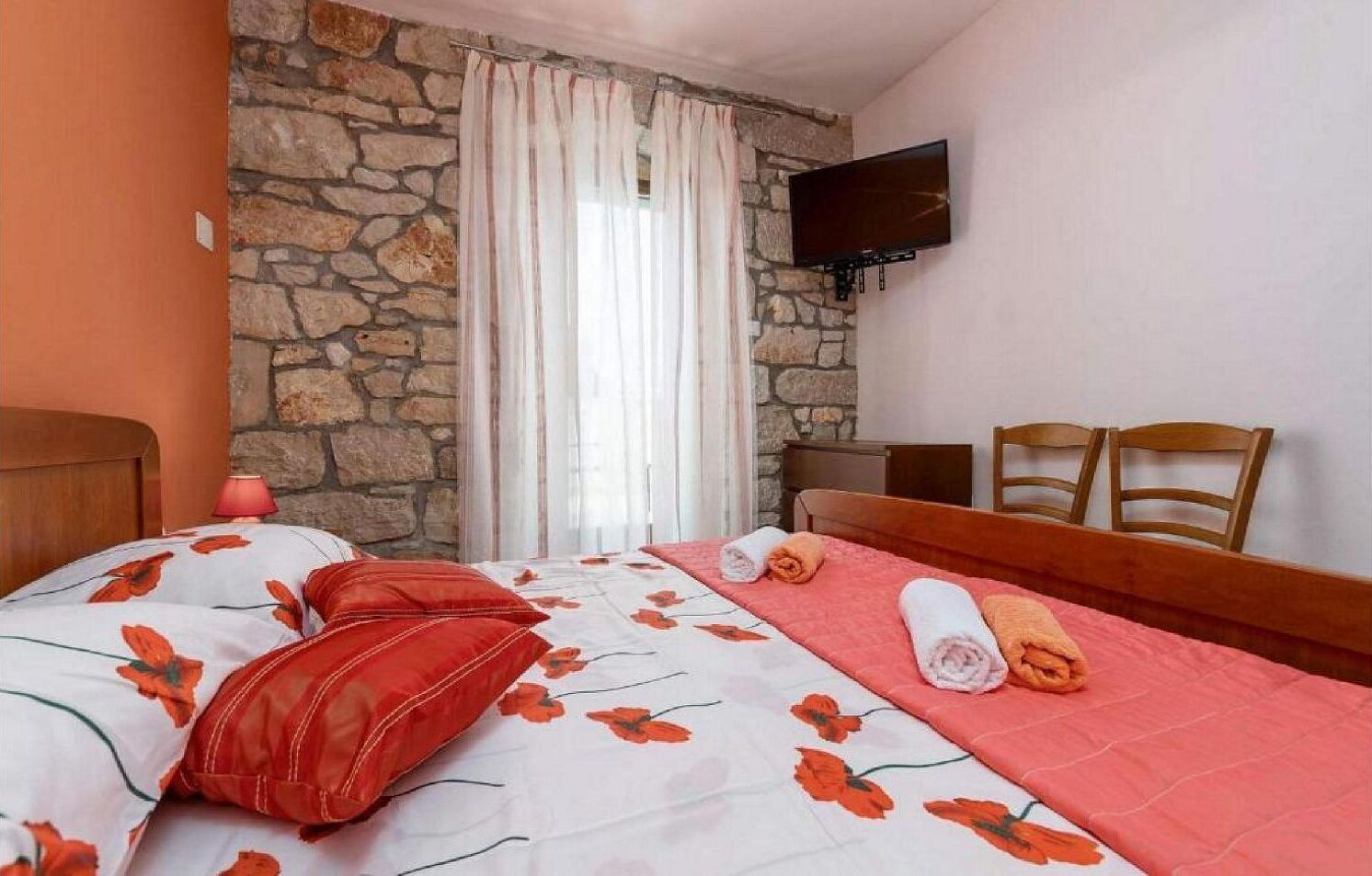 Ein Hotelzimmer mit Steinwand, einem Bett mit floralem Bettbezug, einem an der Wand montierten Fernseher und zwei Holzstühlen.