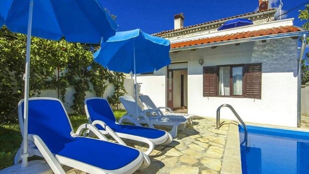 A poolside area with lounge chairs and umbrellas, a stone-tiled patio, and a building with a tiled roof and shutters.
